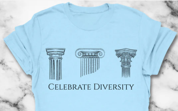 The image shows a light blue t-shirt laid flat on a marble surface. The t-shirt features a graphic design with three classical column capitals, each representing a different style: the Ionic, Corinthian, and Doric orders. These are depicted in a detailed, monochromatic line drawing style. Below the column capitals, the text "CELEBRATE DIVERSITY" is printed in a bold, uppercase font. The overall design emphasizes the theme of diversity through the representation of different architectural styles.
