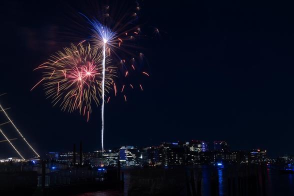 A night scene featuring colorful fireworks in the sky above the Boston city skyline, with reflections in the water below and illuminated buildings in the background.