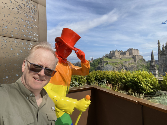 Selfie of the tooter beside the Johnny Walker statue (painted in a Pride rainbow) on a balcony at the Johnny Walker museum and restaurant in Edinburgh. The historic castle is in the background.