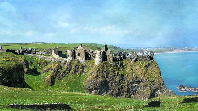 A medieval castle stands on a rugged cliff overlooking a vast expanse of blue sea, with rolling green hills in the background. The structure's stone walls and round towers appear weathered, blending naturally with the landscape. Photography by Debra Martz