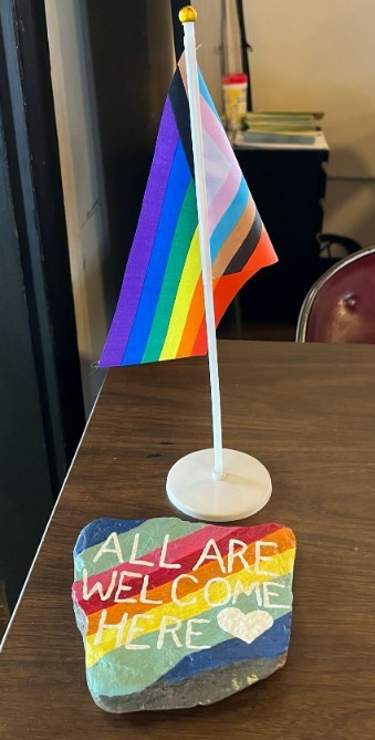 A progress pride flag sits next to a rock that has been painted with rainbow colours and the text "All are welcome here." Picture is from the Midwest Buddhist Temple Bulletin.