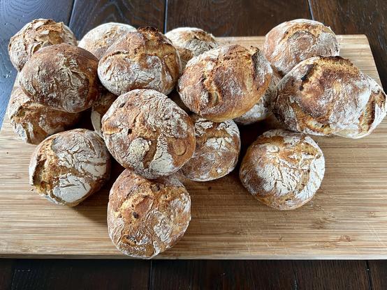 Auf einem dunklen Holztisch liegt ein Holzbrett. Darauf liegen zahlreiche frisch gebackene Brötchen mit rustikalem Ausbund