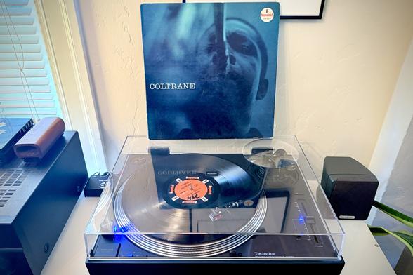 A black vinyl record spins on a black turntable. The record sleeve stands up on the turntable’s closed dust cover behind the platter.