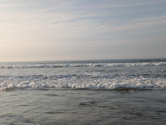 Photo of surf breaking at the ocean's edge. The sky above is streaked with wispy white clouds.