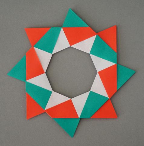 An eight-pointed wreath folded from red and green paper on a grey background. The wreath has rotational symmetry but no mirror symmetry. White triangles surround the central octagonal hole.