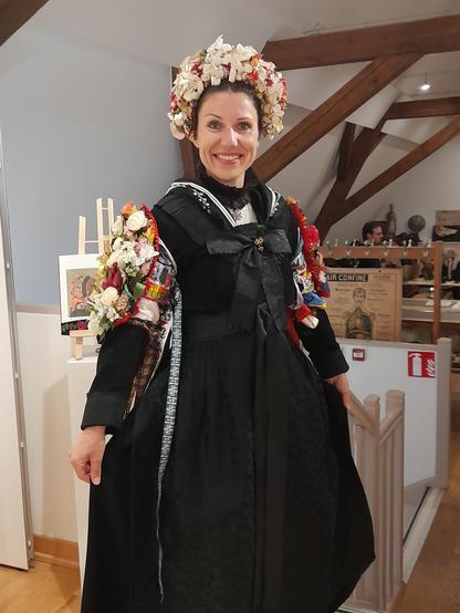 A traditional protestant bride's costume from Northern Alsace, Presented by a woman from the Hunspach folklore group (photographed with permission). The skirt, top and waistcoat, as well as the scarf at the neck are black; silver embroidery was only permitted on the scarf and at the sides. The hair is plaited and pinned up. The bride wears a head-covering ‘crown’ of colourful flowers and glass beads. The same artificial arrangement wraps around the upper arms like a suit of armour. Originally, the flowers were handmade from silk and the beads were mouth-blown. For events, they are replaced by imitations like these made of fabric and coloured plastic, as the real crowns would be too valuable.