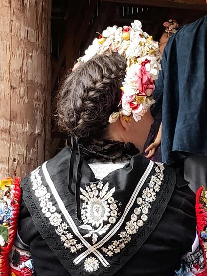The back of the bride's head and shoulders showing the plaited hair and the crown formed like a half very wide headband. The scarf is embroidered with silver - in floral patterns. The traditional patterns like the tree of life, flowers, or ears of grain were symbols for fertility. These scarves were the only decoration for robes  that was allowed for married women by protestants in that region.