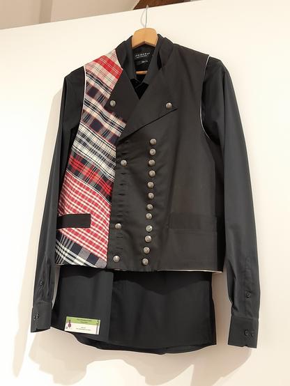 A designer waistcoat that takes up the traditional men's waistcoat shape with a close-fitting parallel row of silver buttons on black and a buttoned lapel. Unlike the old waistcoats, however, the right-hand row of buttons is irregularly reduced to 3 buttons and sthe right side is sewn from red, black and white Kelsch chequered fabric. In older times, Kelsch was reserved for women and household laundry. Crèation by Thibaut Heck.