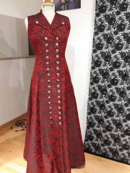 A long designer robe made of a precious fabric with red floral patterns on black ground. It's armless, the top is tight-fitting with a waist, the skirt is wide. Two parallel rows of silver buttons go from the top until the knees, the lapel is buttoned. Pastors would have condemned a woman for such a dress in the 1950s. Because the traditional red fabric and the cut with the buttons were only permitted for men to wear on their waistcoats.