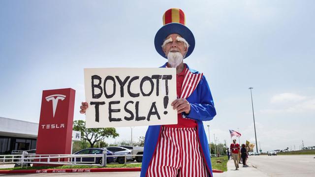 A man dressed as Uncle Sam holds a sign reading 'Boycott Tesla!' outside a Tesla store.