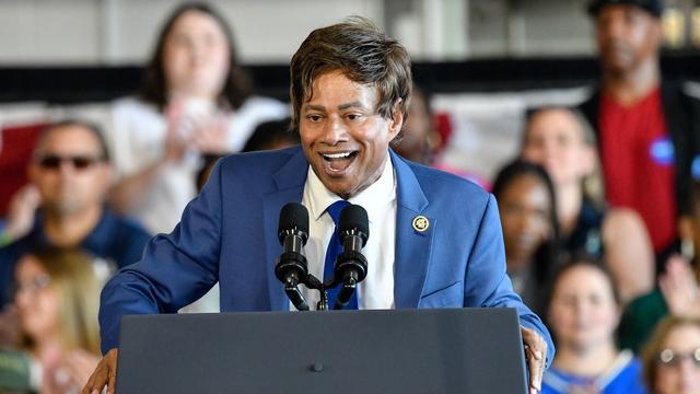 Rep. Shri Thanedar wearing a blue suit and speaking at a gray podium in front of a crowd of people.