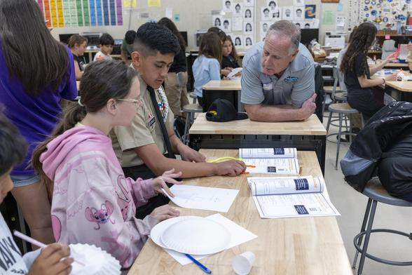 A group of students sitting at a desk in a classroom work on constructing paper airplanes. A man leans on the desk, speaking with the students. Behind the man are other students seated at desks.