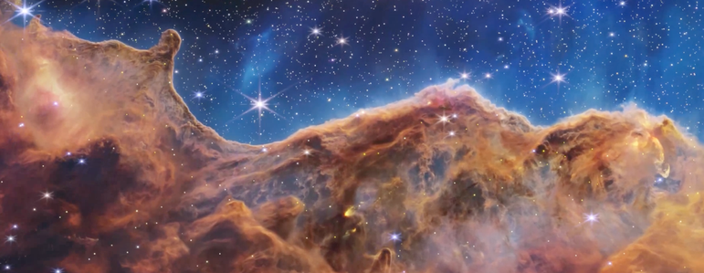 The image is divided horizontally by an undulating line between a brown cloudscape forming a nebula along the bottom and a comparatively clear upper portion in blue. Speckled across both portions is a starfield, showing innumerable stars of many sizes. The upper blue portion has wispy translucent cloud-like streaks rising from the nebula below. The orange and brown cloudy formation in the bottom half varies in density and ranges from translucent to opaque. The nebula contains ridges, peaks, and valleys—an appearance similar to a mountain range. In the bottom left corner, a clearer area free of gas and dust appears black with speckled stars.