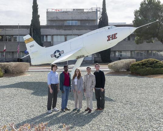 Students pose side-by-side in front of a static aircraft display in front of a building at NASA Armstrong. The students are wearing professional clothes and sneakers. Students pose side-by-side in front of a static aircraft display in front of a building at NASA Armstrong. The students are wearing professional clothes and sneakers.