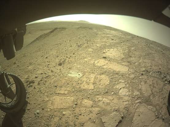 A color image from the Martian surface shows uneven yellow-tan ground, covered in gravel and much larger flat, angular rocks showing above the surface. Parts of the rover are visible but shaded – a portion across the top of the frame, and a wheel in the lower left corner. A drill hole is visible in the ground near the center of the image, surrounded by lighter-colored soil.