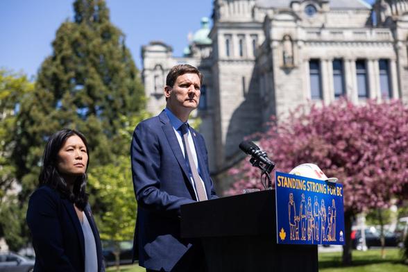 Premier David Eby and Infrastructure Minister Bowinn Ma stand outside the BC legislature. Eby is at a lectern featuring a "Standing Strong for BC sign" with a microphone and a white hard hat on top of it. Ma stands behind him