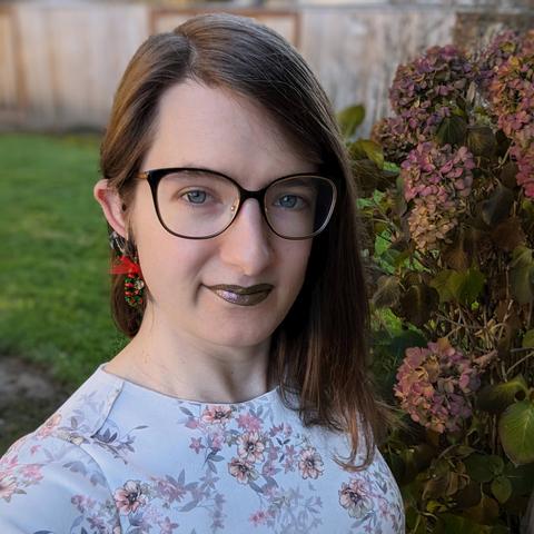 Hazel looks at the camera. She is wearing rose gold lipstick, black glasses, and a cherry blossom dress. In the background are flowers and a fence