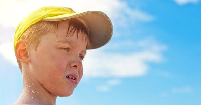 Halbnahe: Ein etwa 6-jähriger Junge mit heller Haut und blonden Haaren im Profil, schaut nach rechts. Er trägt ein gelbes Käppi, Schweißperlen im Gesicht. Im Hintergrund in der Unschärfe Sonne und wenige Wolken.