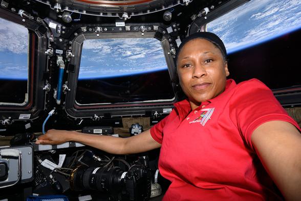 A black woman in a red shirt poses in the International Space Station cupola with the Earth pictured behind her.