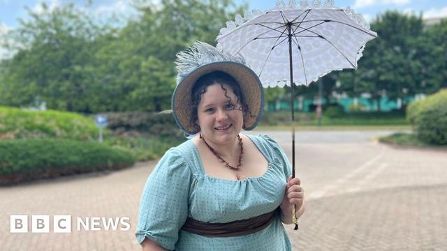 Sophie Andrews stands outside in her regency outfit and looks at the camera, smiling. She has dark brown curly hair, with two strands of ringlets framing her face and the rest of her hair in an up-do underneath a straw bonnet which has a pale blue ribbon rim and blue feathers sticking out the top. Sophie holds a parasol made of white lace. She is wearing a brown stone necklace with a turquoise blue dress, which has three quarter length sleeves and a brown ribbon sash around her waist. The background is blurred but shows trees and hedging surrounding a pale beige cobbled area of road.