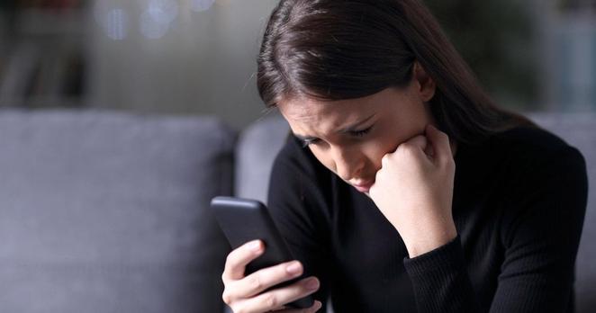 Eine junge Frau mit dunklem langem Haar und dunklem Pullover sitzt auf einer Couch und schaut unglücklich auf ihr Smartphone.