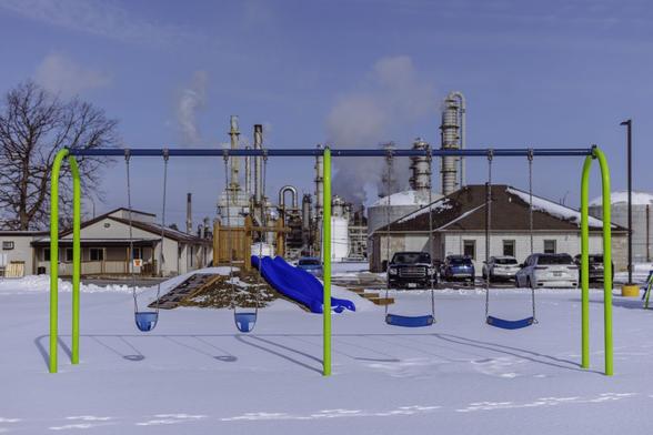 A blue and green swingset in front of a small building, with smokestacks in the background