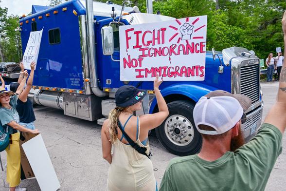 A person holds a sign reading "FIGHT IGNORANCE NOT IMMIGRANTS" at a roadside protest