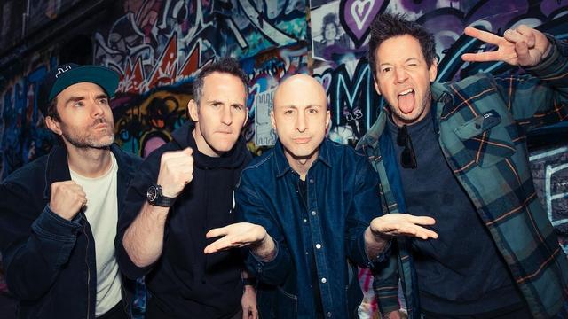 Quatre hommes posent devant un mur plein de graffitis.