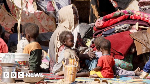 A woman covered by a beige headscarf sits on a blanket, surrounded by five young children in colourful clothing, with braided hair. They are sitting amid piles of fabric, clothing and matting, under makeshift tents that have been propped up by small trees.