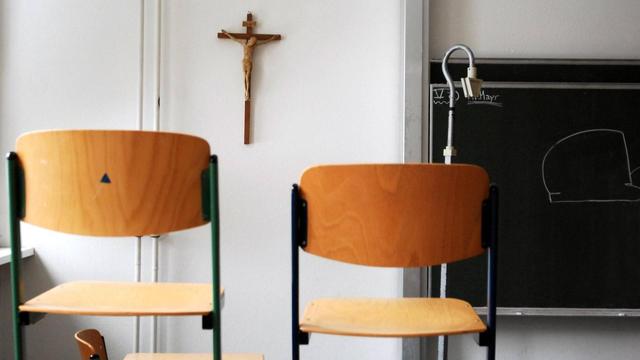 Hochgestellte Stühle in einem Klassenzimmer - im Hintergrund an der Wand ein Kruzifix.