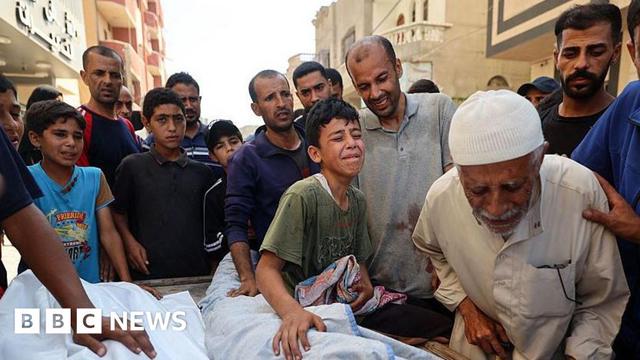 A group of men and boys stand around a child in a body bag, some crying and touching the shroud, during a funeral on the street in Gaza.