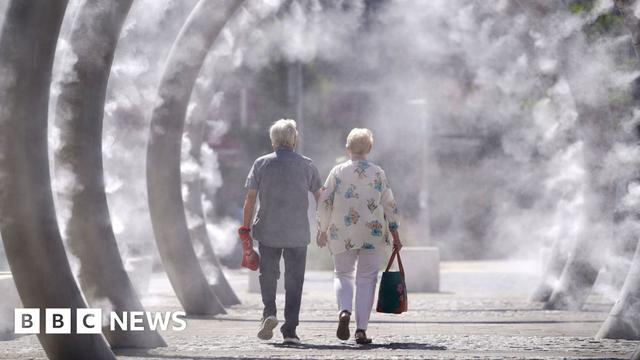 A man and a woman - both with silver hair - are walking inside a row of ring metal structures that are spraying mist on a very sunny day. A man is wearing a checkered blue shirt with short sleeves, jeans and trainers and a woman in white trousers and a loose-fitting shirt.