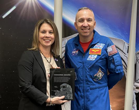 A woman in business casual attire receives a plaque from a male astronaut in a blue flight suit.