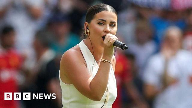 Claudia Rose singing before the match. She is wearing a cream halter neck waistcoat and has dark hair with a central parting tied back in a bun and chunky gold earrings. The crowd is blurred in the background