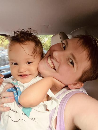A light-skinned biracial baby with dark curly hair smiles slightly and holds their hand up to the side of their parent's face. The parent, who is white, has their head tilted to the side with a big smile, and has short light brown hair. The background is the inside of a vehicle.