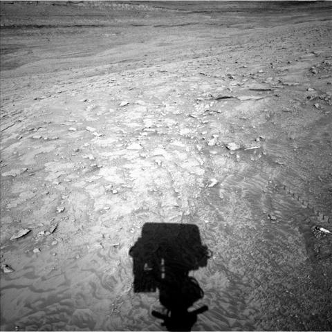 A grayscale photo from the Martian surface shows a rough, rocky landscape in medium gray, with a shadow of the Curiosity rover’s boxy masthead showing at the bottom center of the frame.