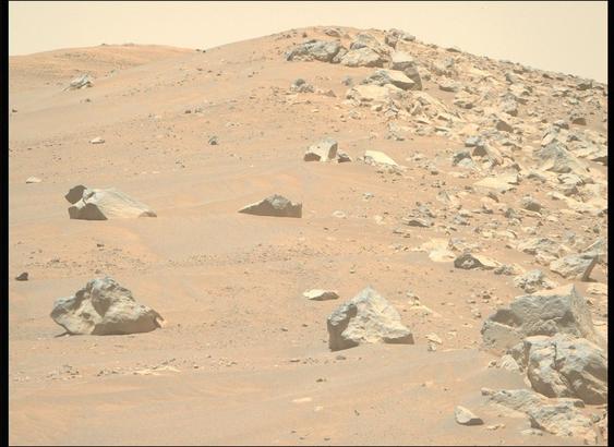 A color photo from the Martian surface shows a landscape of sandy, rocky terrain. What looks like a pair of light orange-brown sand dunes rise in the background. Four or five large, sharp-edged rocks are strewn among the sand on the left side of the image. But a clear vertical dividing line extends from the center of the image to the right side, where the sand is covered by similar rocks of all sizes. A pale yellow sky is visible above the scene.