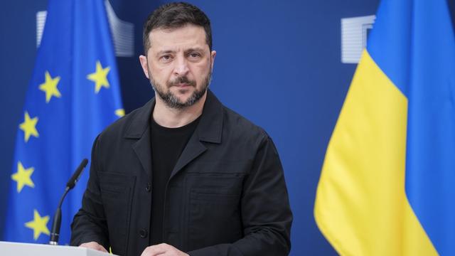 A man with short dark hair and beard stands behind a white podium with a microphone, flanked by blue EU and yellow-blue Ukrainian flags on a blue background. A man with short dark hair and beard stands behind a white podium with a microphone, flanked by blue EU and yellow-blue Ukrainian flags on a blue background.