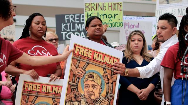 A diverse group of people at a protest holding signs saying "FREE KILMAR" and "BRING HOME Kilmar" with an illustrated portrait of a man behind bars in red, blue, and orange colors.
