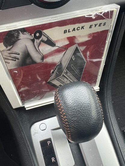 CD case for the Dischord Records album Black Eyes. Red and white cover with a human male singing into a microphone with a tape recorder below it. The cover has very constructivist design scheme to it. The pic is set up in my Subaru. 