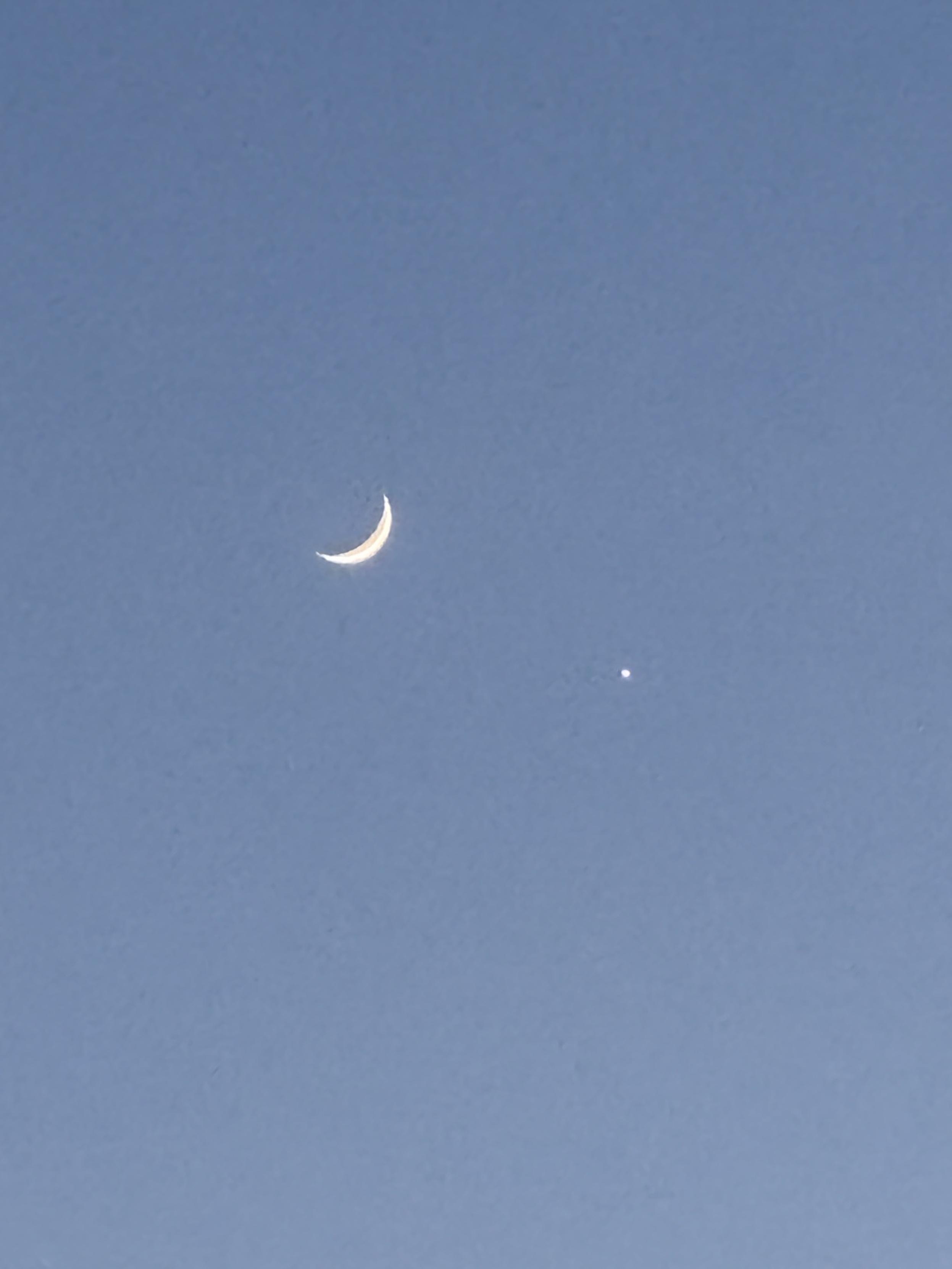Slim, thin crescent moon in a pale blue twilight hour sky with Jupiter very close by.